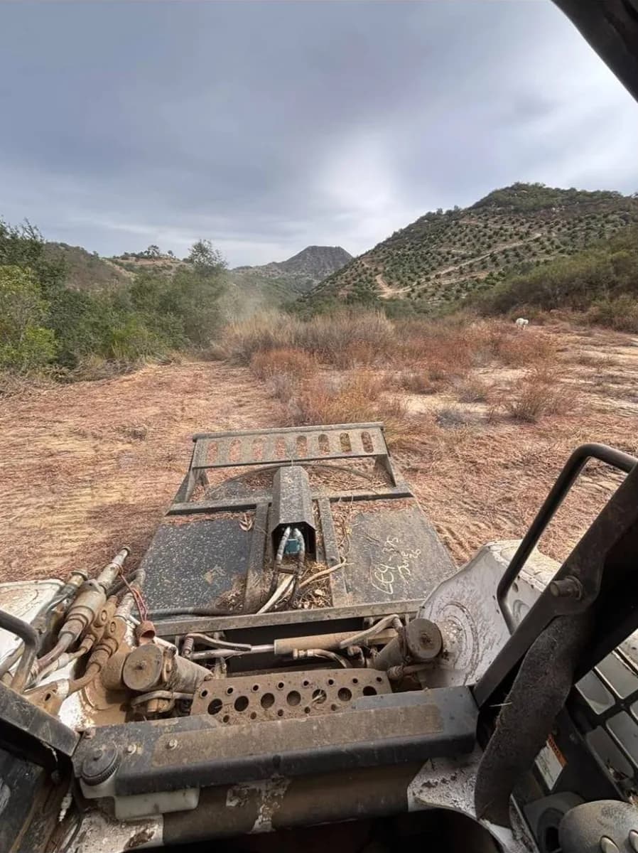 Brush cutter on the front of the Bobcat T300 facing a field of dry brush with terraced avocado groves on the hills beyond