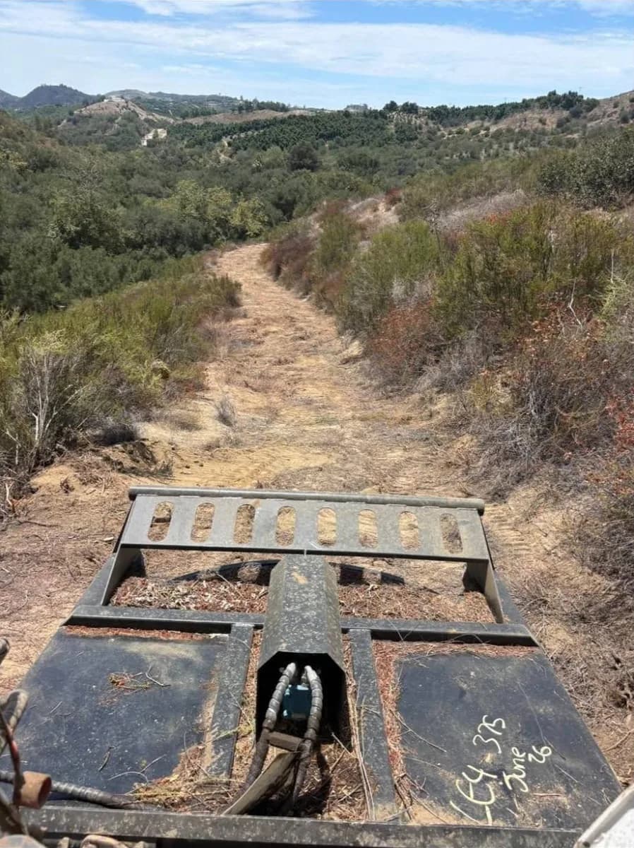 Cleared fire break trail running downhill through chaparral toward avocado groves, seen over the brush cutter