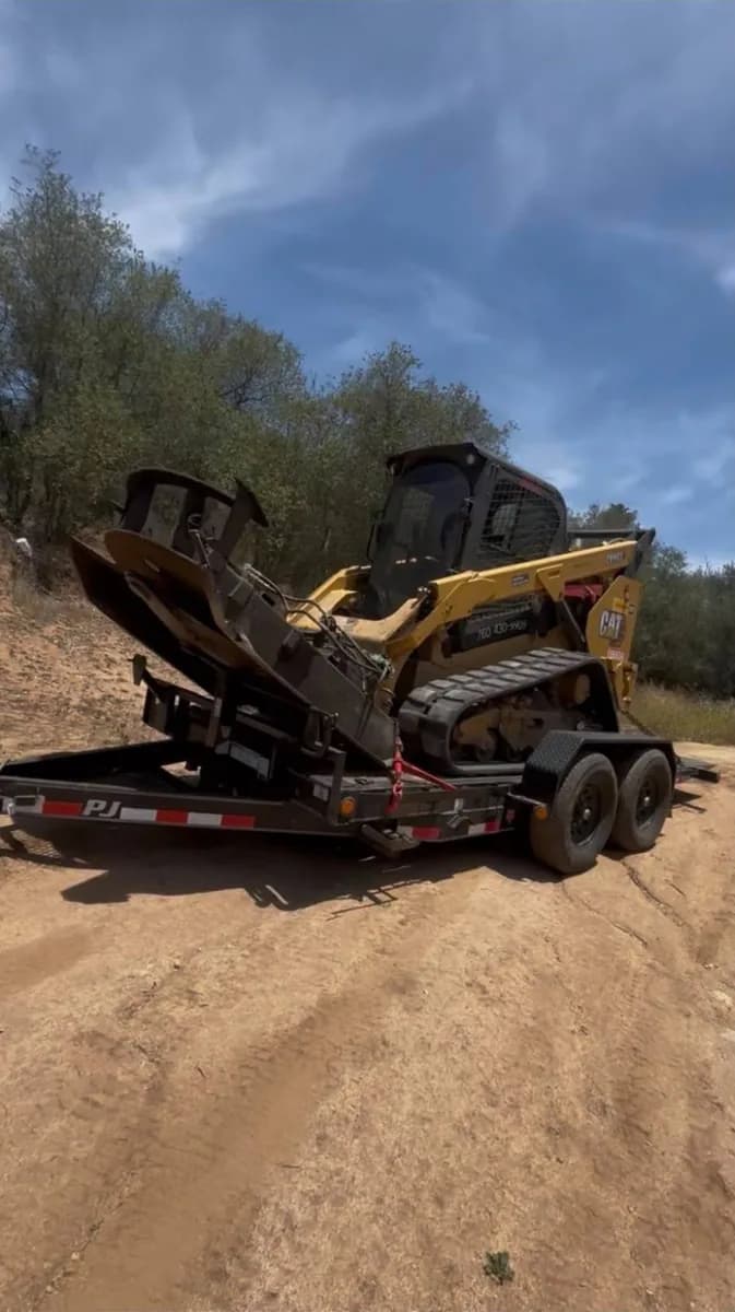 Tracked loader with a brush grapple chained down on a tilt trailer on a dirt road